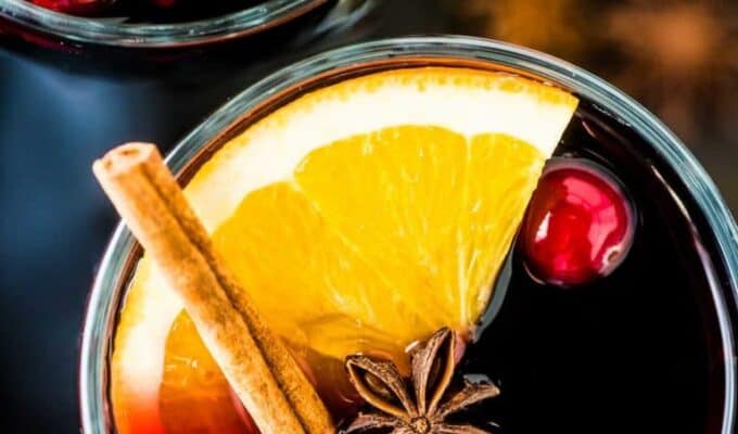 A close up of a glass of mulled wine with an orange slice, cinnamon stick, and a few cranberries in it.