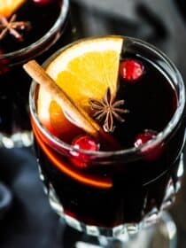 A glass of mulled wine with an orange slice, cinnamon stick, and a few cranberries in it.