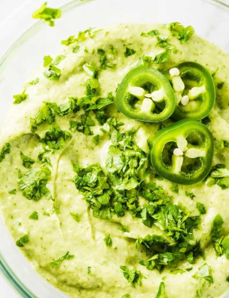 Creamy Avocado Sauce in a glass bowl with minced cilantro and sliced of jalapeño peppers on top.
