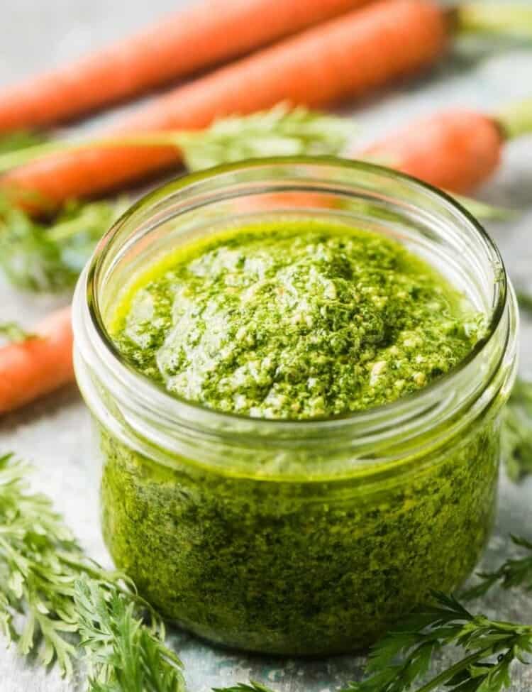A glass jar filled with carrot top pesto with some orange carrots and carrot greens behind it.