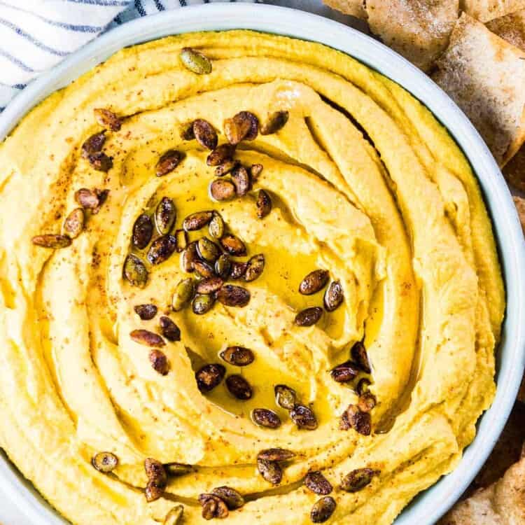 A blue bowl filled with pumpkin hummus with homemade pita chips surrounding it.