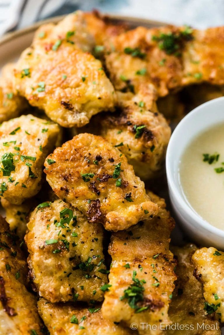 Healthy grain free chicken nuggets piled on a plate with a tiny bit of parsley on top and dipping sauce at the side. 