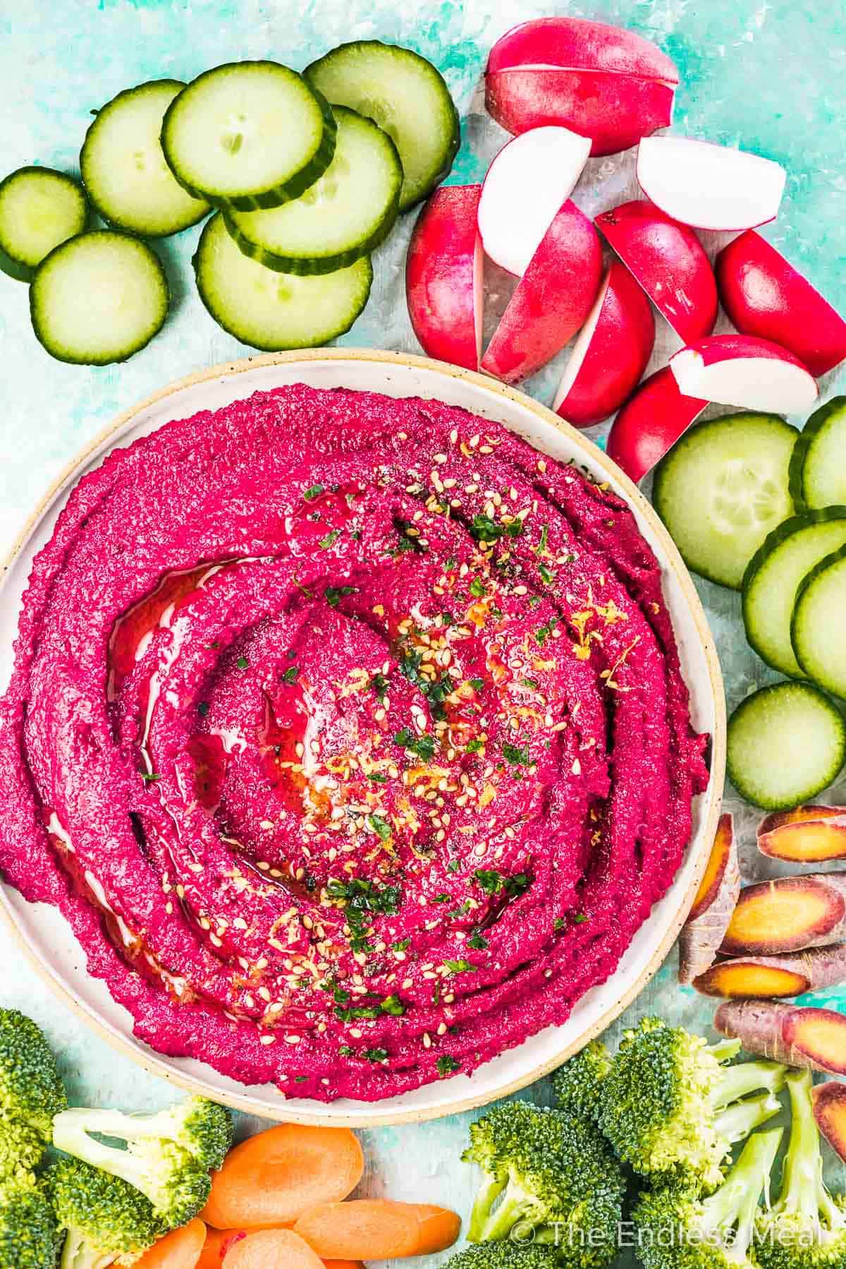 Beet hummus in a white bowl surrounded by veggies.
