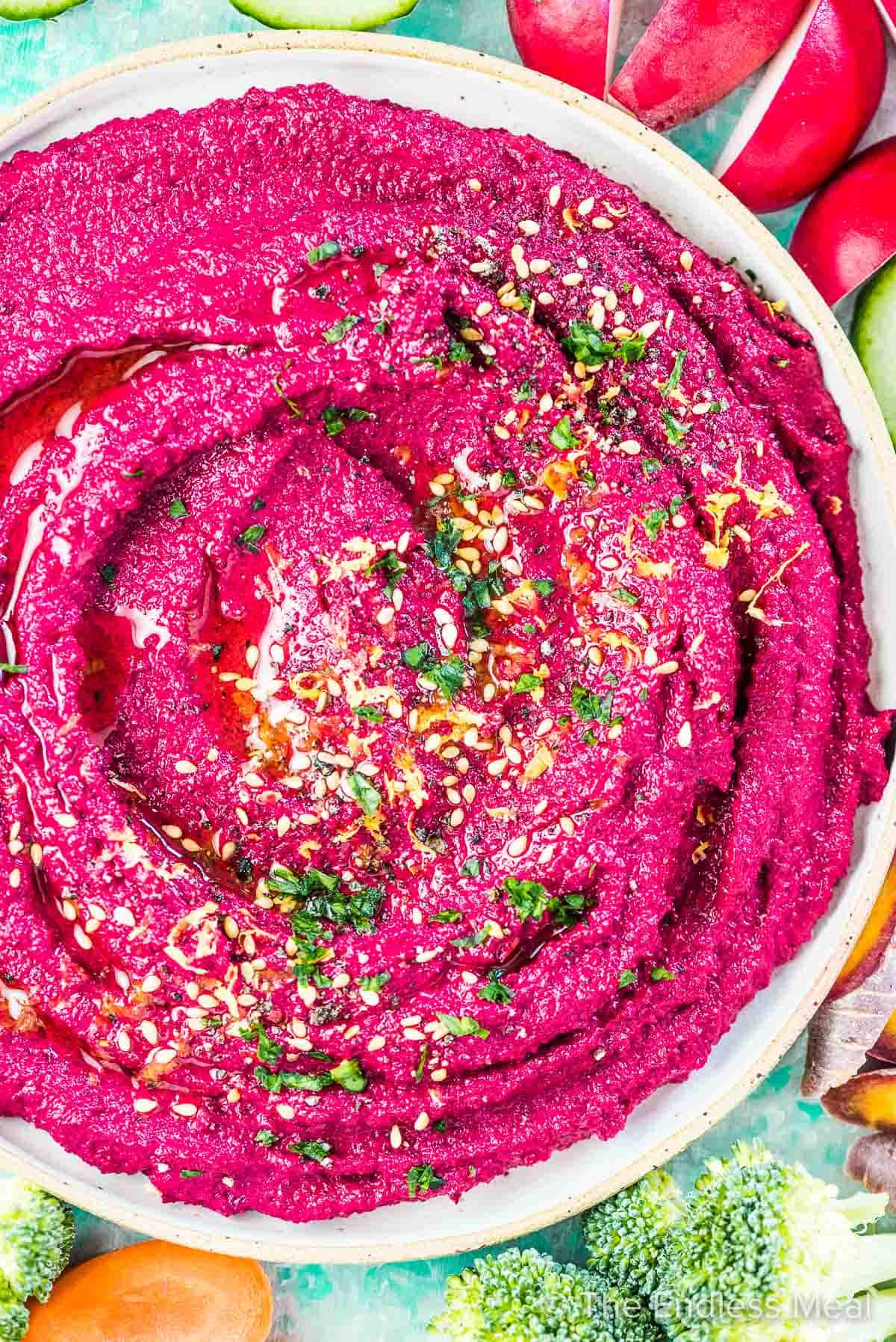 A close up of beet hummus in a white bowl surrounded by veggies.