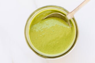 A metal spoon rests in a jar filled with creamy green goddess dressing, viewed from above on a white surface.