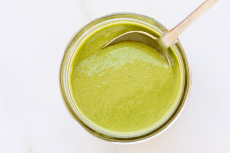 A metal spoon rests in a jar filled with creamy green goddess dressing, viewed from above on a white surface.
