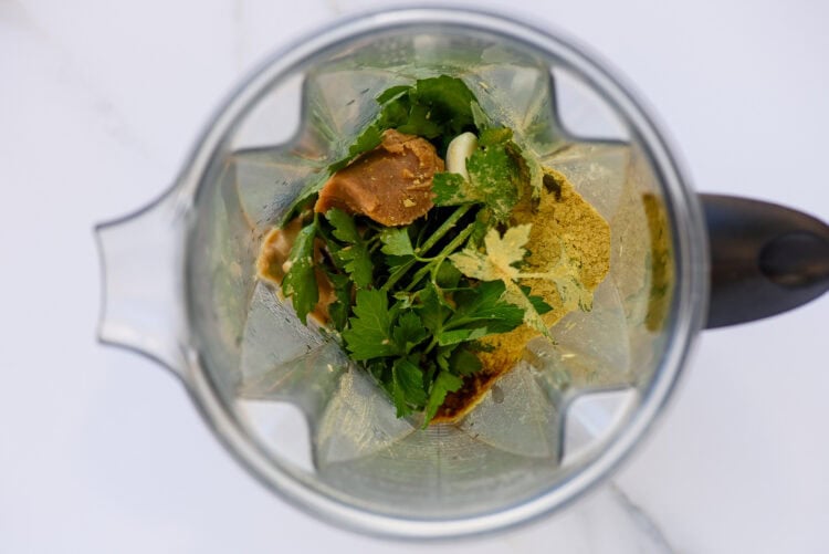 Overhead view of a blender containing fresh parsley, nutritional yeast, a brown paste, and other ingredients on a white surface—perfect for making homemade green goddess dressing.