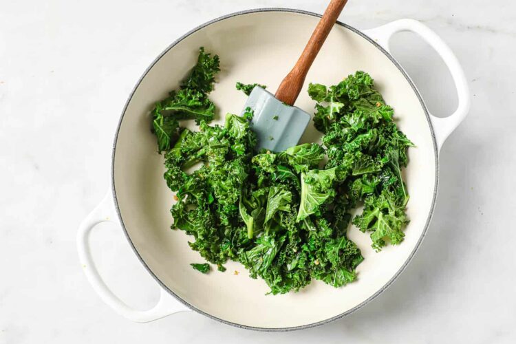 Chopped kale being sautéed in a white pan with a spatula on a light surface, perfect for adding to dairy-free pumpkin alfredo.