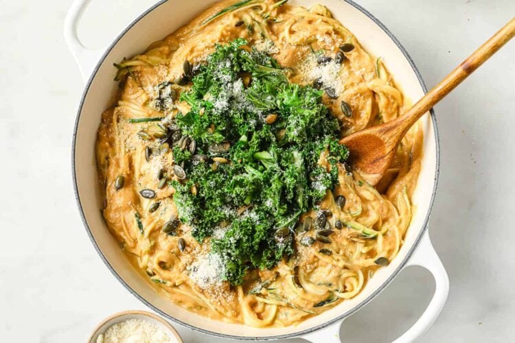 A pot of creamy dairy-free pumpkin alfredo pasta topped with chopped kale, seeds, and grated cheese, with a wooden spoon resting inside.