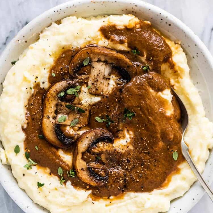 A plate of mashed potatoes covered with vegan mushroom gravy and a couple of mushrooms on the top.