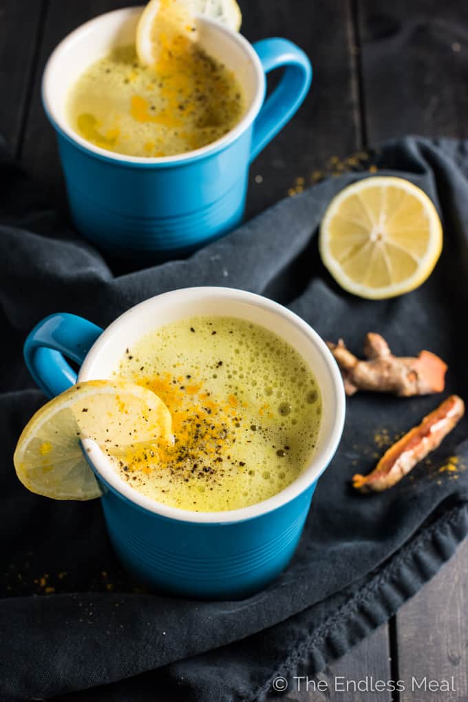 Mugs of turmeric tea with fresh turmeric slices and lemons beside them.
