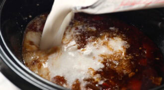 Coconut Milk is being poured from a measuring cup into a slow cooker filled with a brown liquid mixture.
