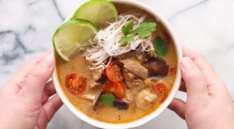 A bowl of thai chicken noodle soup with sliced cherry tomatoes, mushrooms, chicken, cilantro, and lime wedges, held by two hands on a marble surface.