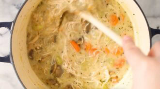 A hand stirs a pot of creamy turkey mushroom soup with shredded chicken, carrots, celery, and mushrooms using a wooden spoon.