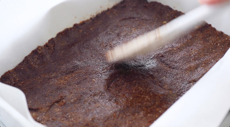 A hand uses a spatula to smooth a thick layer of chocolate batter in a parchment-lined baking dish.