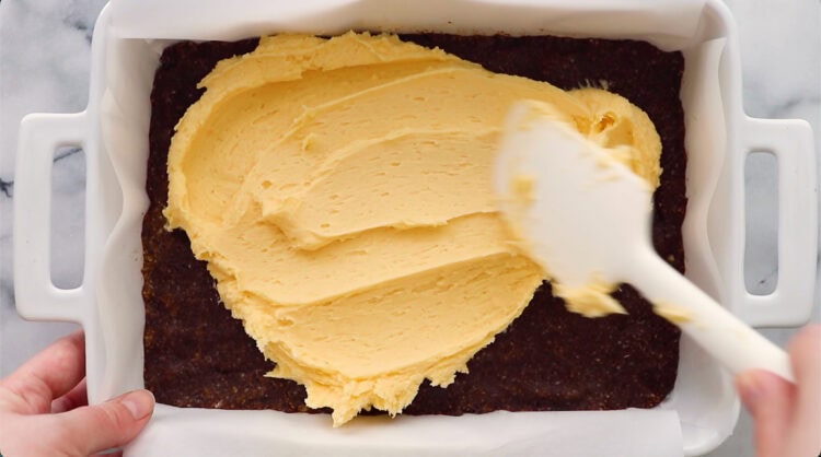 A hand spreads yellow frosting over a dark brown crust in a rectangular white baking dish using a white spatula.