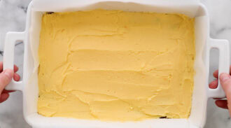 A white rectangular baking dish filled with an even layer of yellow batter, held by two hands on opposite handles.
