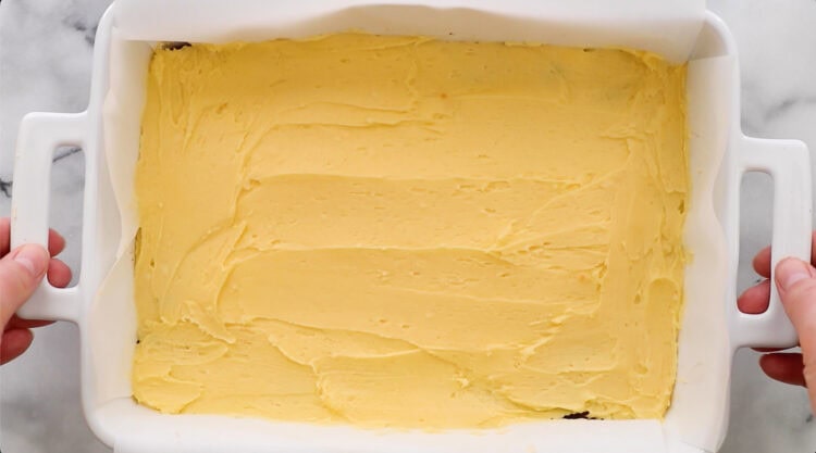 A white rectangular baking dish filled with an even layer of yellow batter, held by two hands on opposite handles.