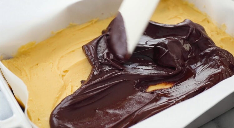 A spatula spreads a layer of chocolate over a base of peanut butter mixture in a parchment-lined baking dish.