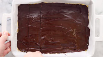 A rectangular pan of dessert with a shiny chocolate topping is being sliced into squares.