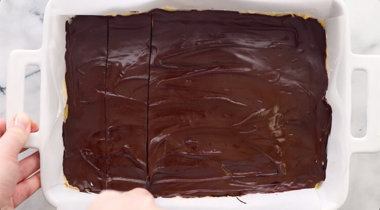 A rectangular pan of dessert with a shiny chocolate topping is being sliced into squares.