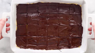 A white baking dish with neatly cut chocolate-topped bars, viewed from above. Two hands hold the dish by its handles.