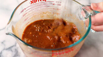 A hand stirs a reddish-brown liquid with chunks in a Pyrex measuring cup on a marble surface.