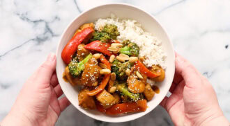 A bowl of white rice with stir-fried chicken, broccoli, red bell peppers, peanuts, and sesame seeds, held by two hands over a marble surface.