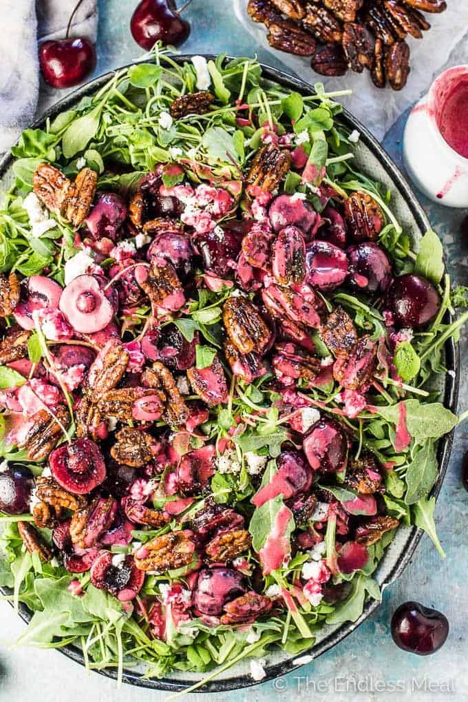 A platter of arugula and fresh cherry salad on a blue wooden table with fresh cherries scattered around.