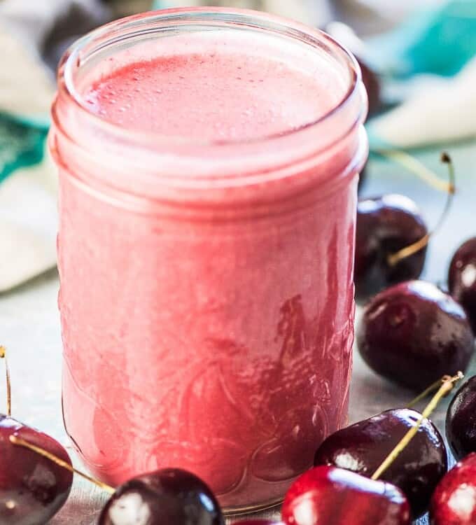 A glass jar full of fresh cherry vinaigrette ready to be poured on the cherry salad. It is sitting on a blue table with fresh cherries around the jar.