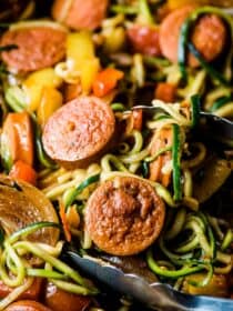 A close up picture of sausage and peppers in a pan with zoodles and a pair of kitchen tongs.