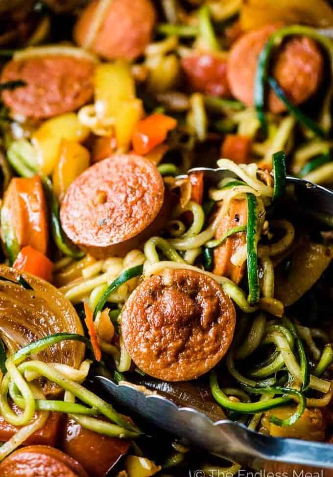 A close up picture of sausage and peppers in a pan with zoodles and a pair of kitchen tongs.