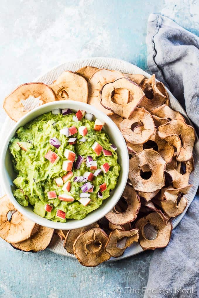 A bowl of apple guacamole with Martin's Apple Chips on the side on a blue and white wooden table. 