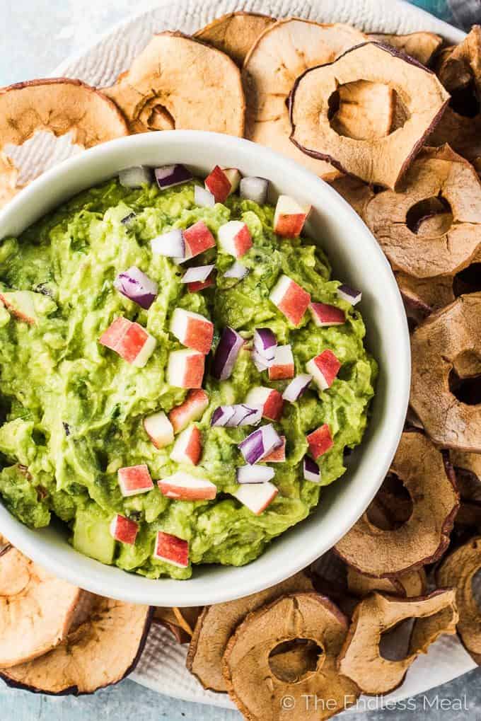 A bowl filled with apple guacamole with minced red apples and red onions on top and apple chips on the side for dipping.
