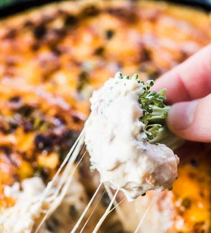 A hand dipping a piece of broccoli in the hot cheese dip.