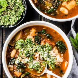 Two bowls of the best minestrone soup recipe on a black table with a bowl of basil pesto on one side.