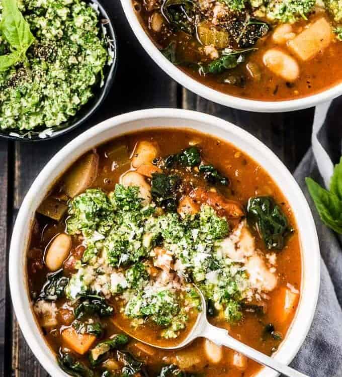 Two bowls of the best minestrone soup recipe on a black table with a bowl of basil pesto on one side.
