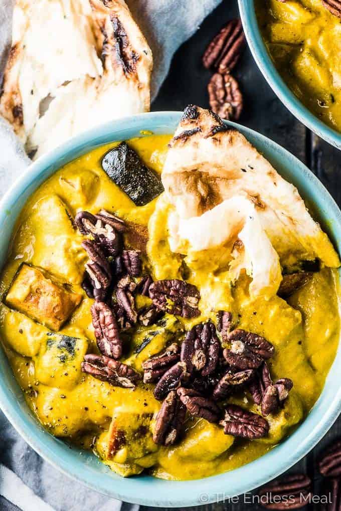 A blue bowl filled with bright yellow pumpkin curry topped with pecans and a piece of naan bread.