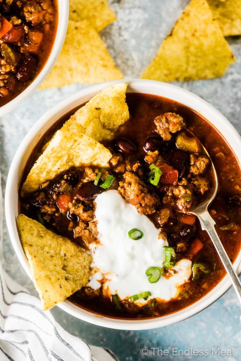 A bowl of healthy turkey chili with sour cream and green onions on top and a few tortilla chips and a spoon. 