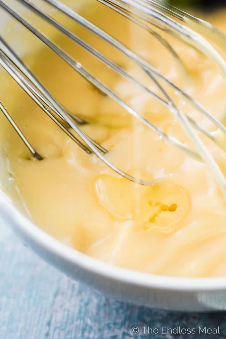 Oil very slowly being poured into a bowl of homemade mayonnaise with a whisk in it. 