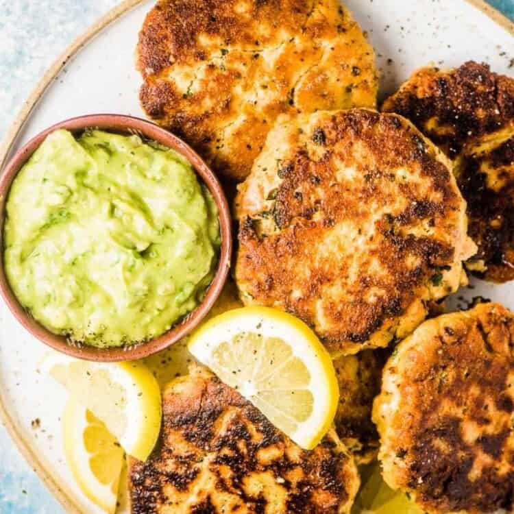 All the fish cakes from this salmon cakes recipe on a white plate with lemons and avocado tartare sauce on the side.