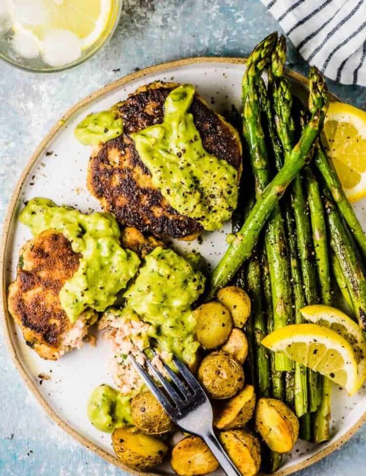 A dinner plate with salmon cakes topped with avocado tartare sauce and asparagus and roast potatoes on the side.