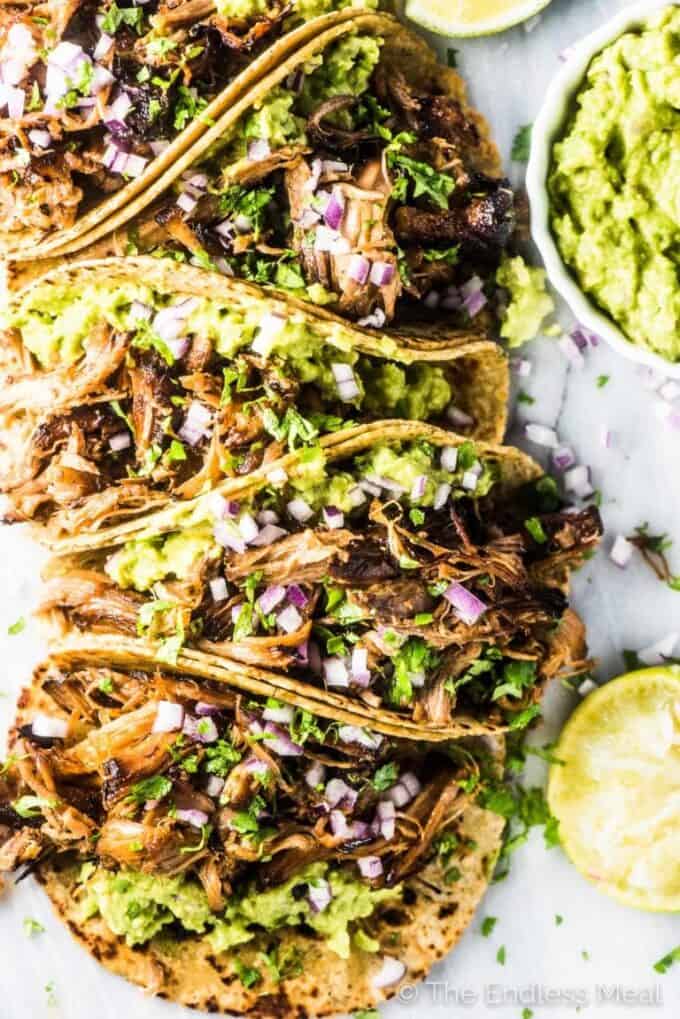 Carnitas tacos with guacamole and red onions with a bowl of guacamole beside the tacos.