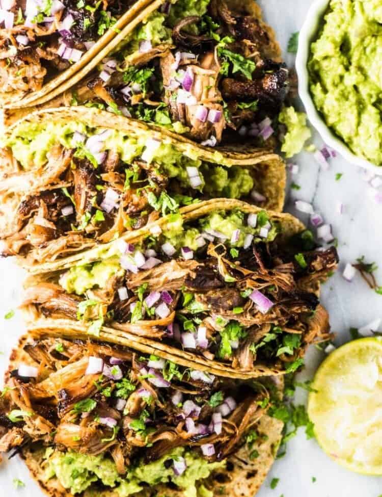 Carnitas tacos with guacamole and red onions with a bowl of guacamole beside the tacos.
