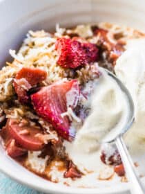 A close up of a spoon in a bowl of strawberry rhubarb cobbler with some vanilla ice cream.