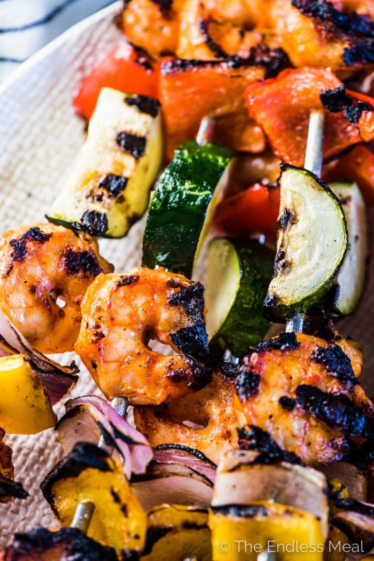 A closeup of grilled shrimp kebabs with summer veggies on a white plate. 