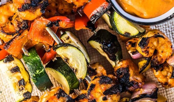 Grilled shrimp skewers on a white plate with veggies and roasted red pepper dip.