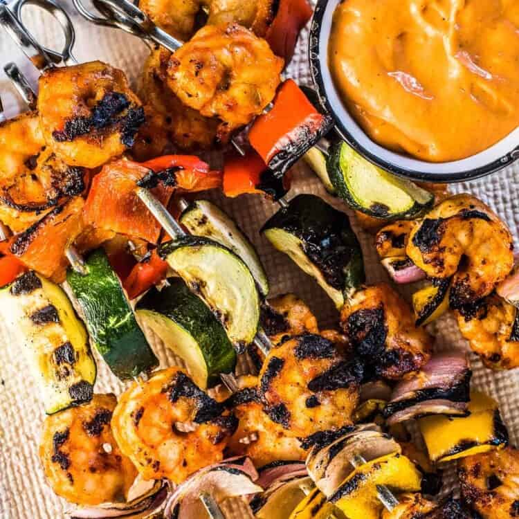 Grilled shrimp skewers on a white plate with veggies and roasted red pepper dip.