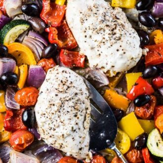 Two tzatziki chicken breasts on a baking sheet surrounded by roasted Greek veggies.