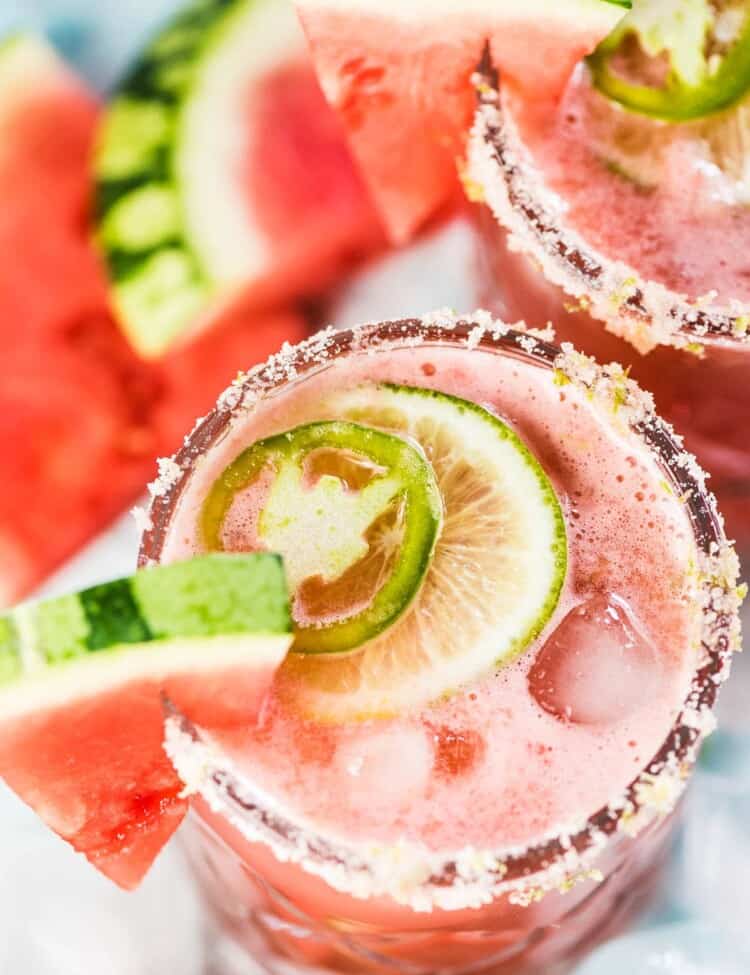 2 glasses of watermelon margarita with a watermelon slice on the side and a lime and jalapeno slice on top.